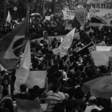 Imagem em preto e branco mostra centenas de pessoas em manifesta₵ao no Rio de Janeiro. Há uma bandeira do Brasil no centro da imagem e bandeiras de movimentos sociais empunhadas por manifestantes. Ao fundo, é possível ver policiais em uma barreira.