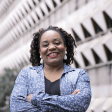 A foto mostra Priscila Cristina Evangelista, uma mulher negra de cabelo castanho escuro cacheado à altura do ombro. Ela veste uma blusa preta com uma camisa azul e branca por cima, brincos brancos e batom vermelho. Priscila está de braços cruzados, sorrindo, em primeiro plano. Ao fundo, a imagem mostra uma parede branca e algumas árvores